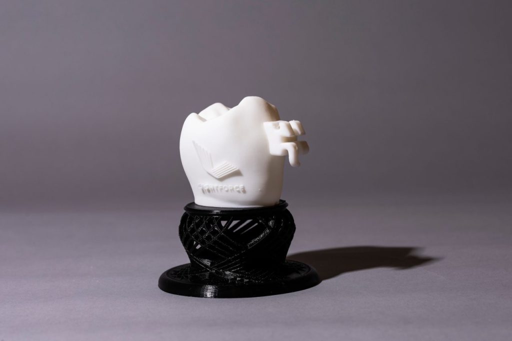Wide angle shot of a 3D Printed ceramic model of a tooth with a lightforce logo embossed on it. The model has a mounted white colored LightForce braces bracket. The model is resting atop a 3d printed table.